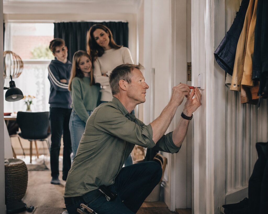 A man installs a device by a doorway while his family watches from the background with curiosity and mild skepticism.
